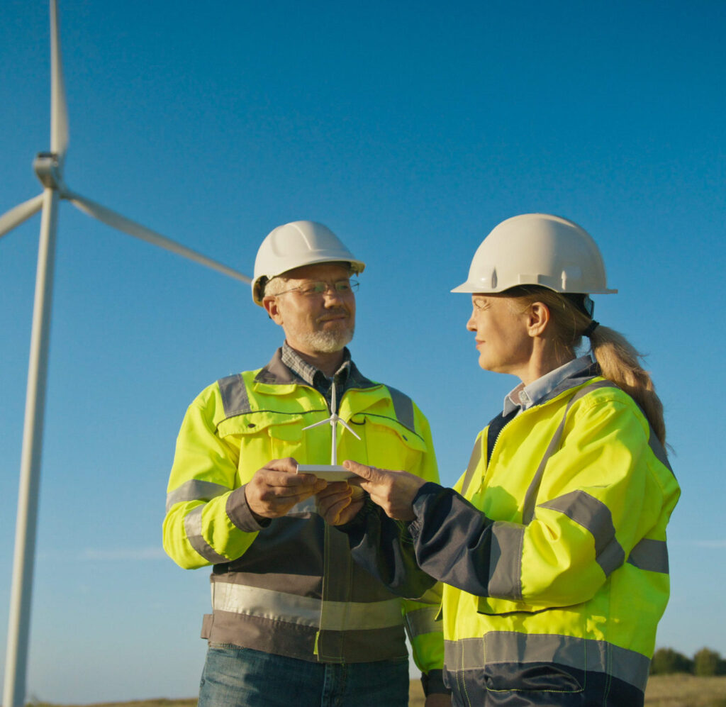Two engineers in hard hats and safety jackets holding a model wind turbine at a wind farm