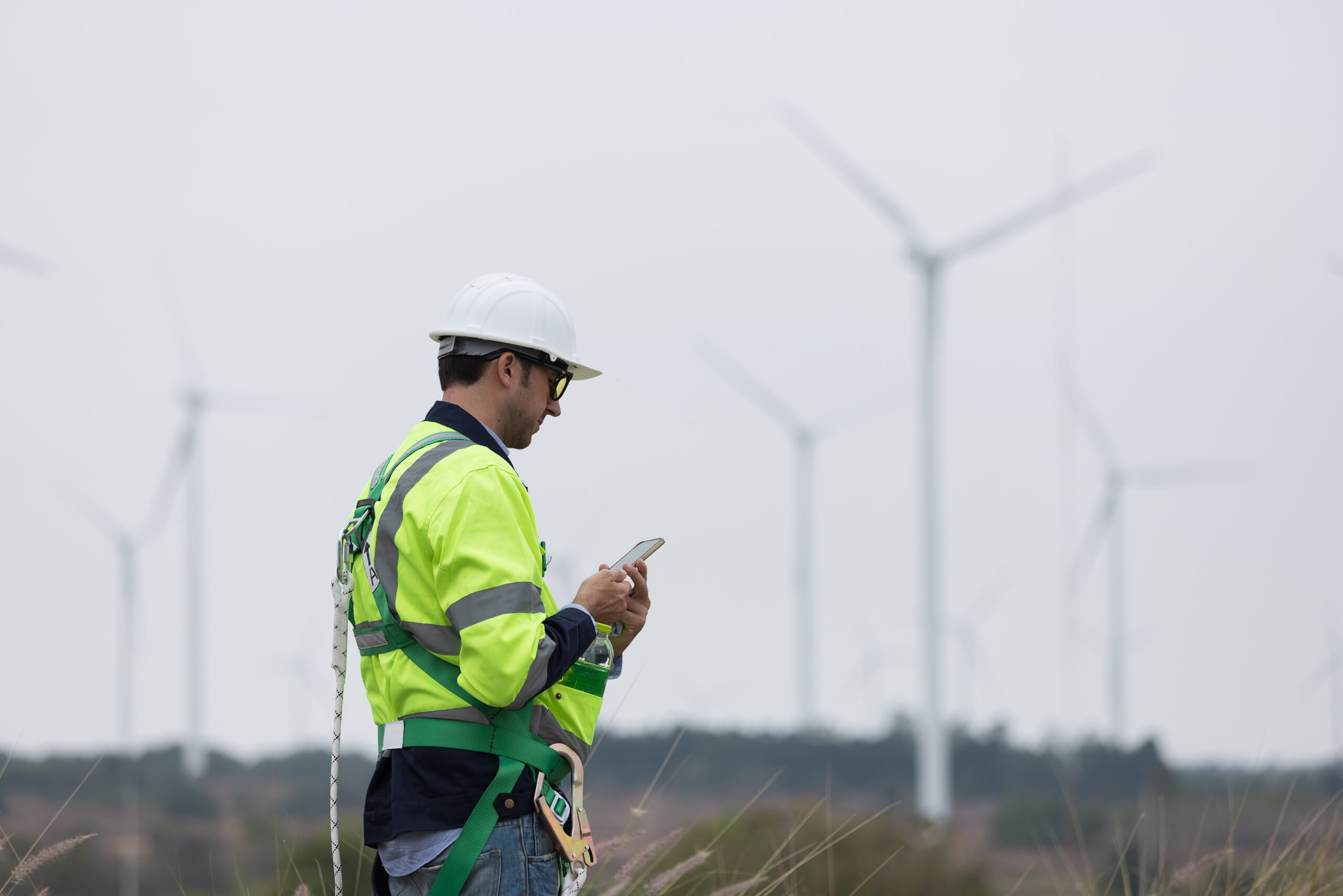 Engineer in a hard hat and safety harness checking a phone at a wind farm