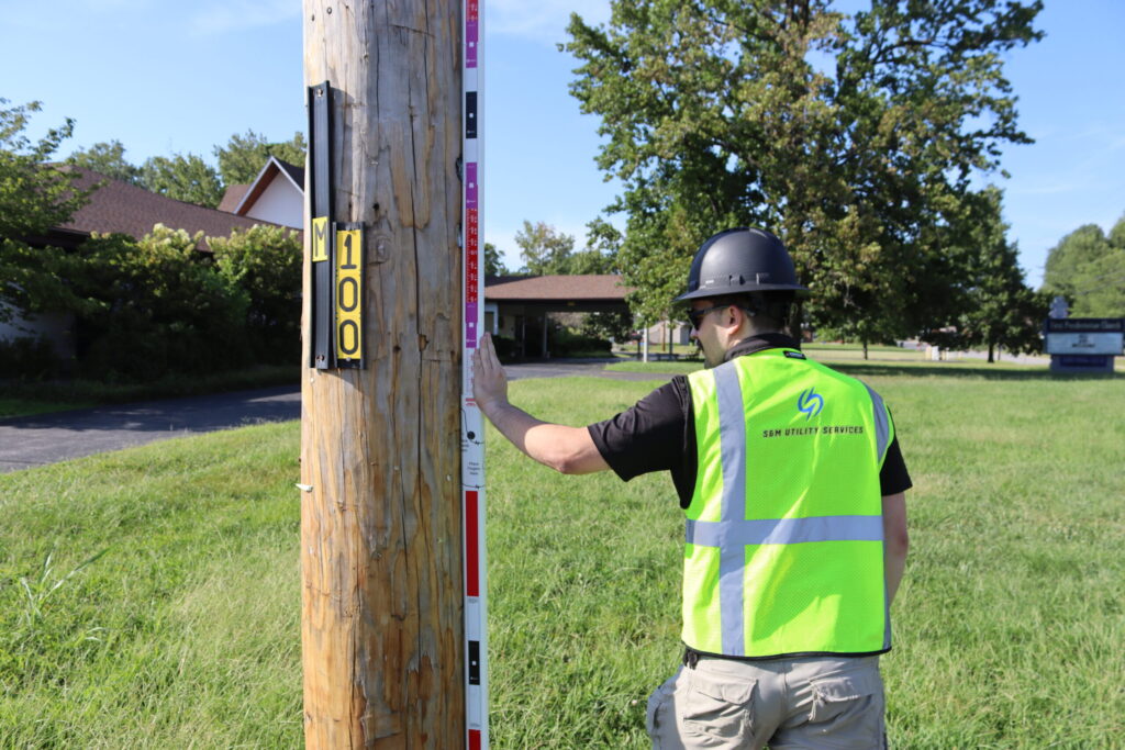 S&M field technician in a hard hat and safety vest measuring a wooden utility pole