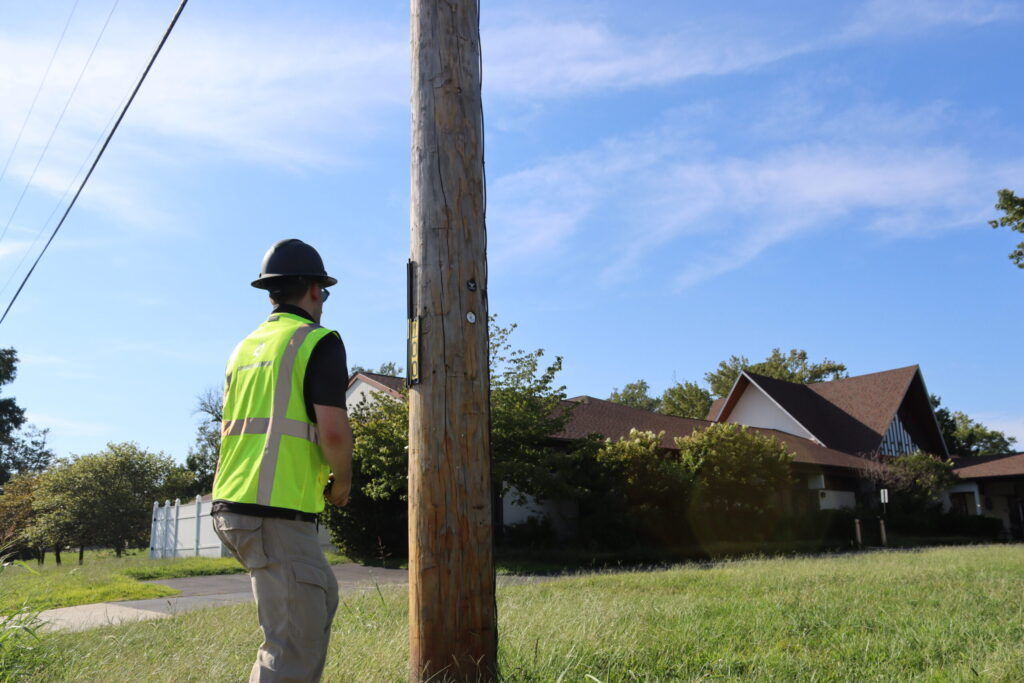 Field technician in a hard hat and safety vest inspecting a utility pole in a neighborhood