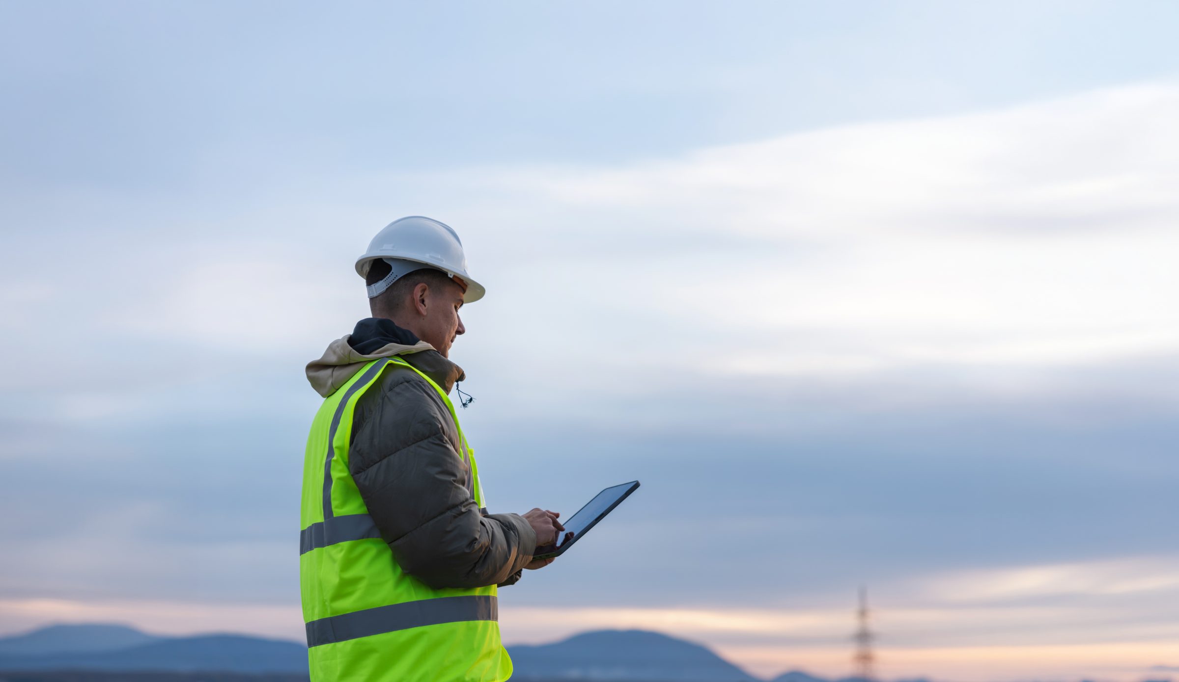 Engineer in a hard hat and safety vest reviewing a tablet at dusk