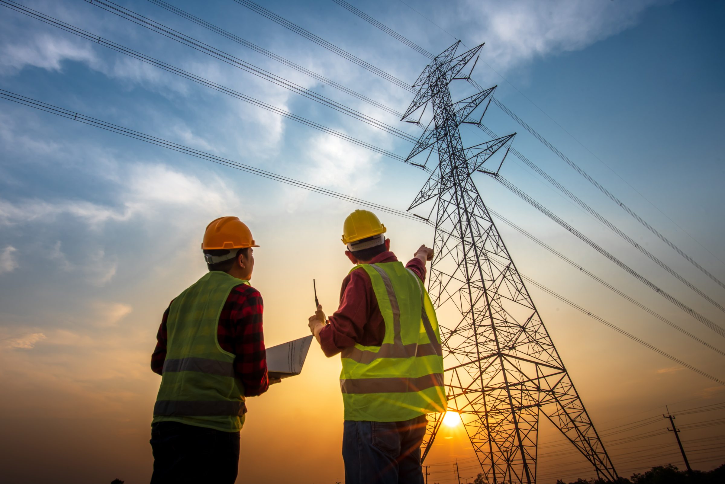 Two engineers in hard hats pointing at a transmission tower at sunset